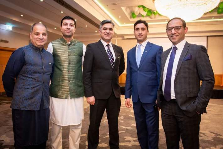 A group of five men in formal and traditional attire at a community event in a banquet hall.