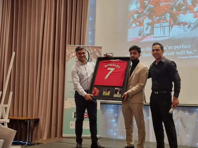 Three men on stage holding a framed Manchester United jersey with a tribute to Cristiano Ronaldo on screen behind them.