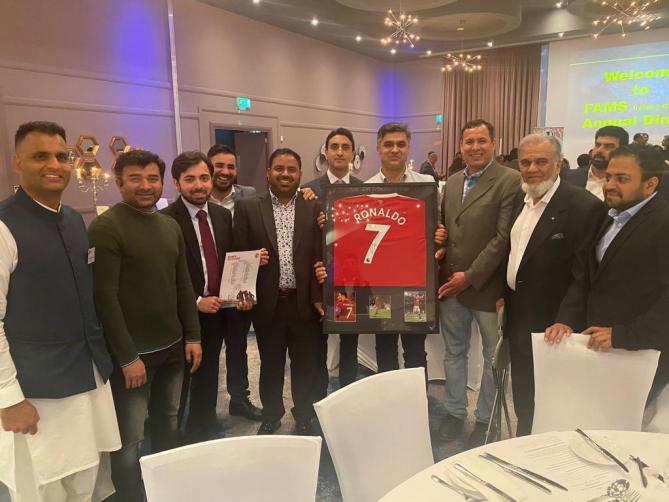 A large group of men at a formal dinner holding a framed Manchester United jersey; welcome screen for an annual dinner in the background.