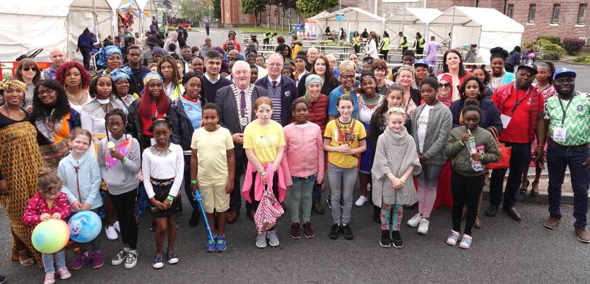 A large diverse group at an outdoor multicultural community festival with tents and a civic official in a chain of office.