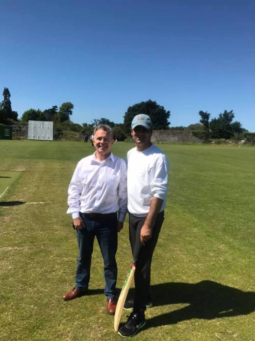 Two men smiling on a green cricket pitch; one holds a cricket bat under a bright blue sky.