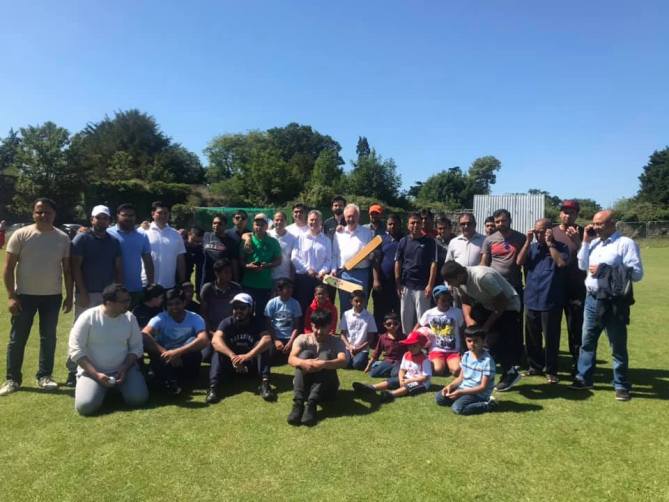 A large group of men and children posing on a cricket field on a sunny day, some holding cricket bats.